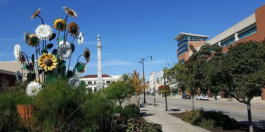Large metal flower sculpture on a street in downtown Racine, Wisconsin with a monument in the background.