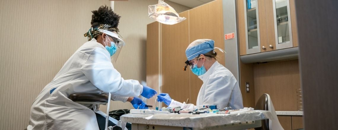 A dentist and dental assistant wearing protective gear work with instruments in a clinical setting.