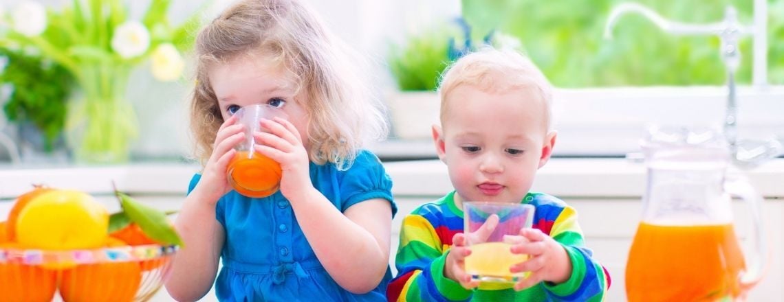A young girl and toddler boy drinking fresh orange juice at a kitchen table.