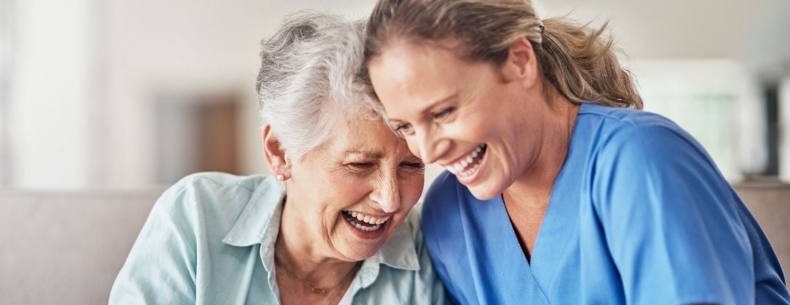 A happy senior woman and her friendly caregiver in blue scrubs share a warm laugh together.