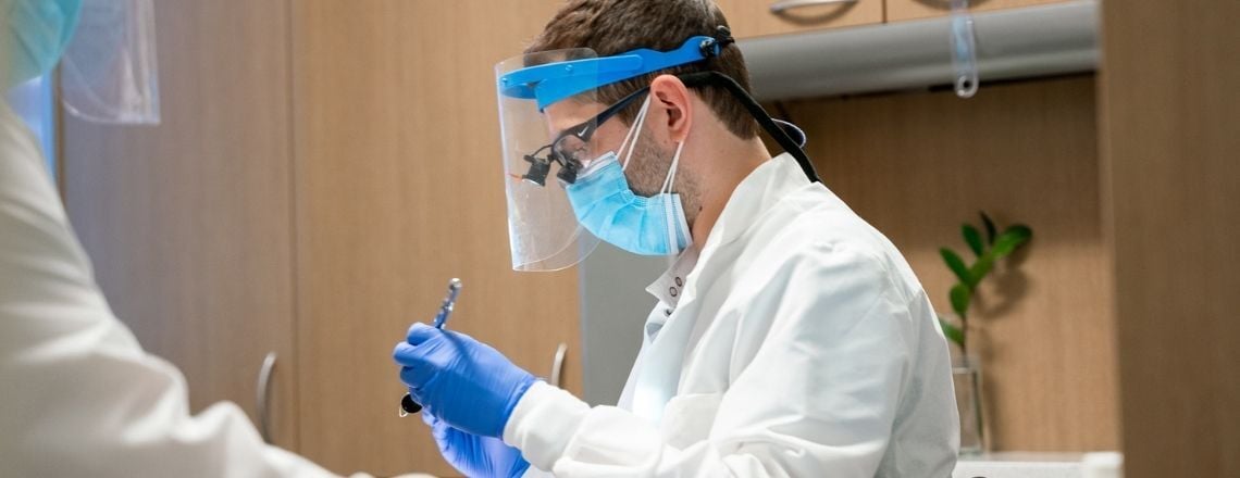 Dentist wearing protective face shield and surgical mask prepares a dental instrument for a procedure.