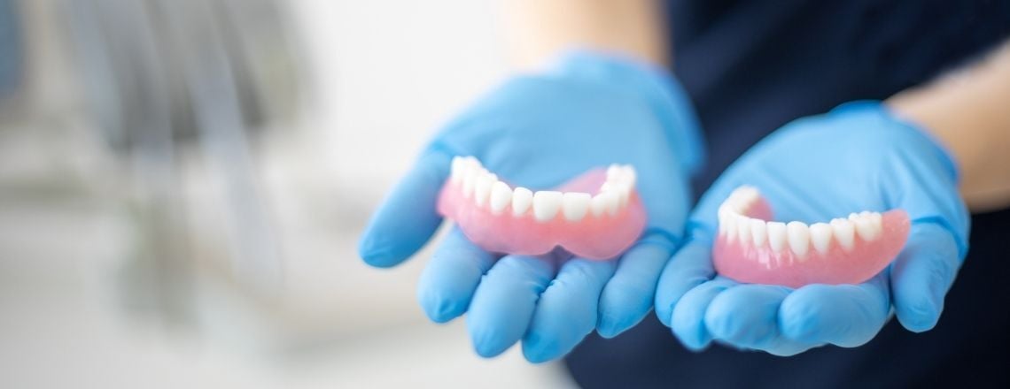 A dental professional wearing blue gloves holds a complete set of upper and lower dentures.