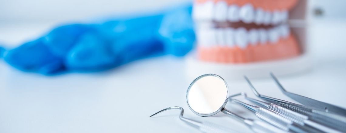 Dental examination instruments including a mouth mirror and probe with a teeth model in the background.