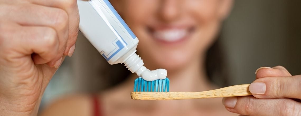 A person squeezing white toothpaste onto a bamboo toothbrush with blue bristles.