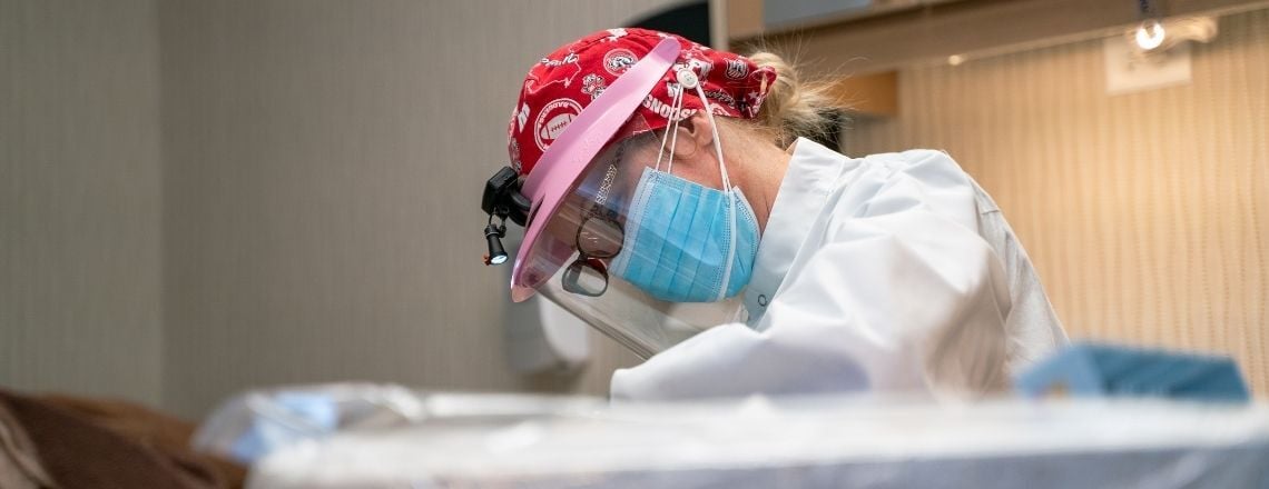 A dentist wearing full PPE, including a face shield and headlamp, performs a dental procedure.