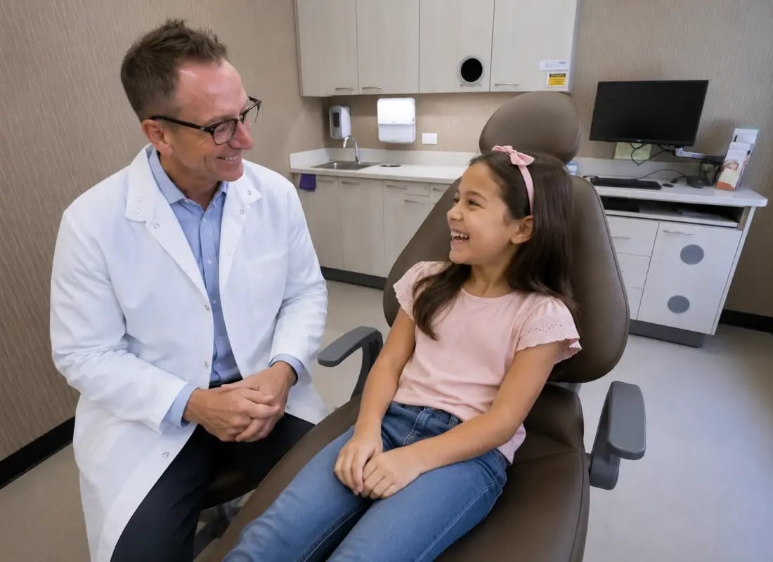 A friendly pediatric dentist smiling at a happy young girl sitting in a dental chair during her checkup.