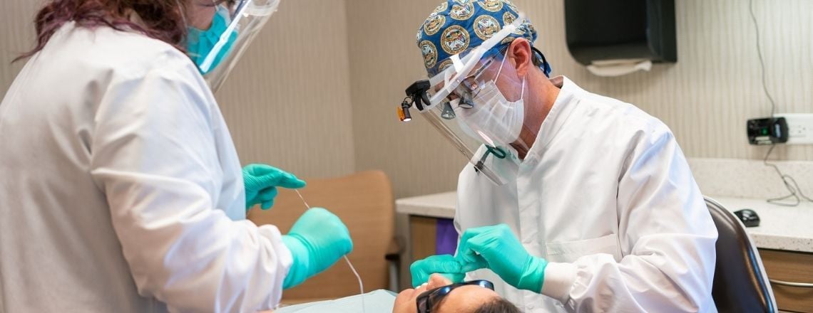 A dentist and assistant wearing protective gear perform a dental procedure on a patient.