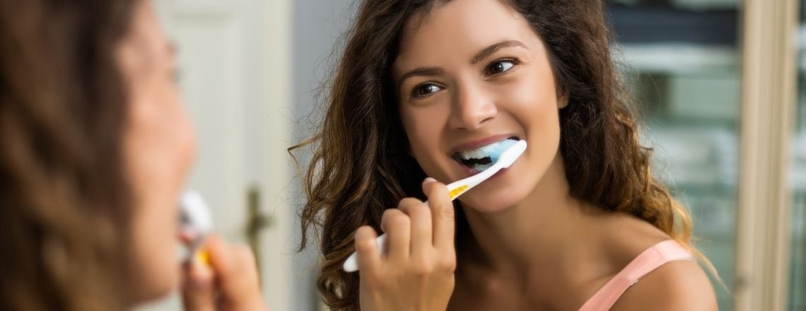 A smiling young woman brushing her teeth while looking in a bathroom mirror.