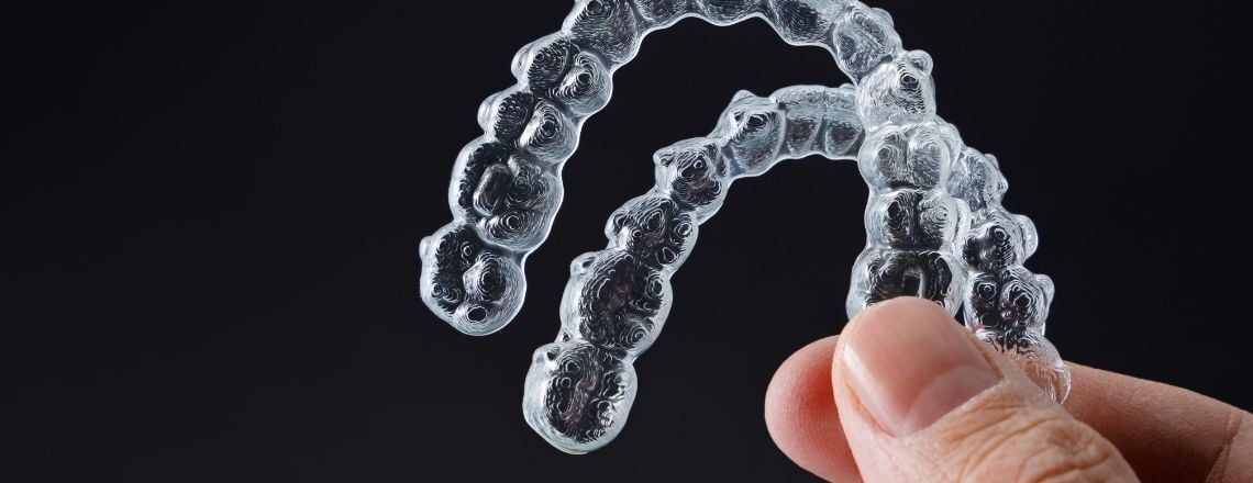 A hand holding a pair of clear plastic orthodontic aligners against a dark background.