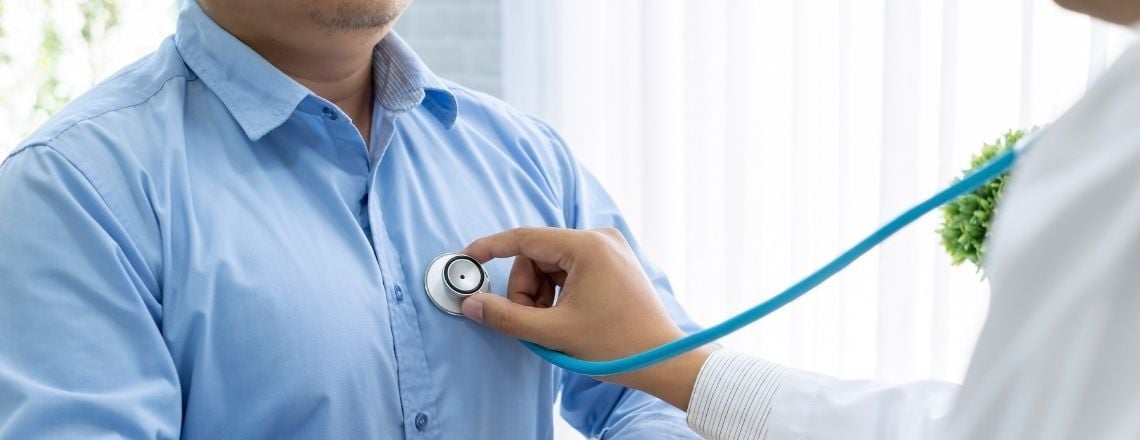 A doctor uses a blue stethoscope to listen to a male patient's heart during a medical checkup.