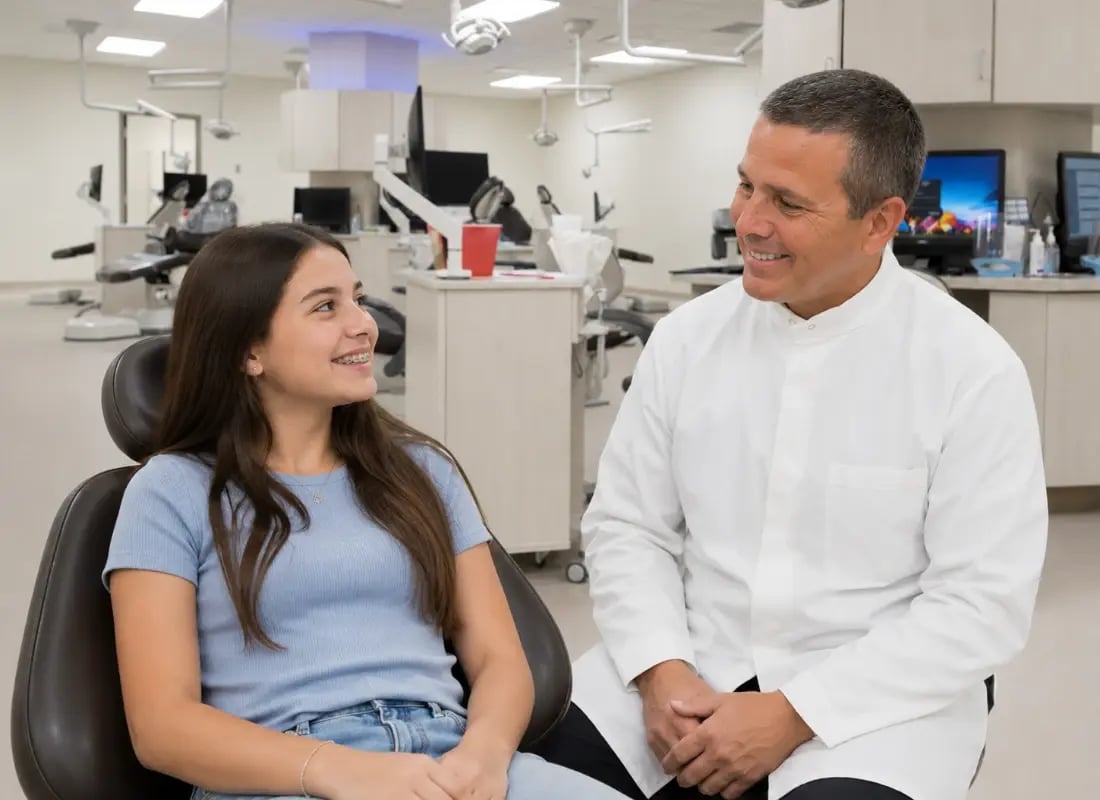 A friendly male orthodontist smiling at a young teenage girl with braces in a dental clinic chair.