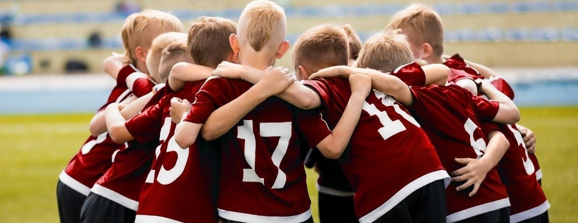 A youth sports team wearing red uniforms huddles together on a grassy field.