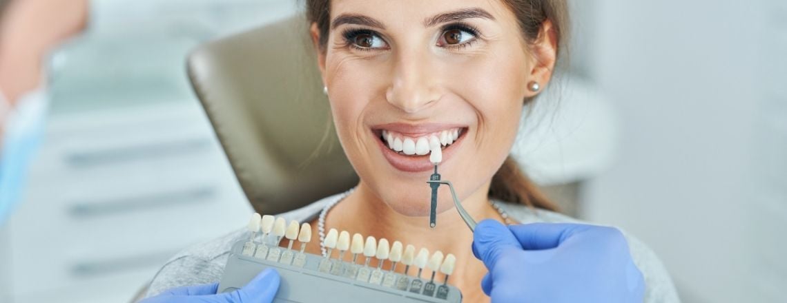 Dentist matching a smiling female patient's tooth shade using a dental color guide chart.