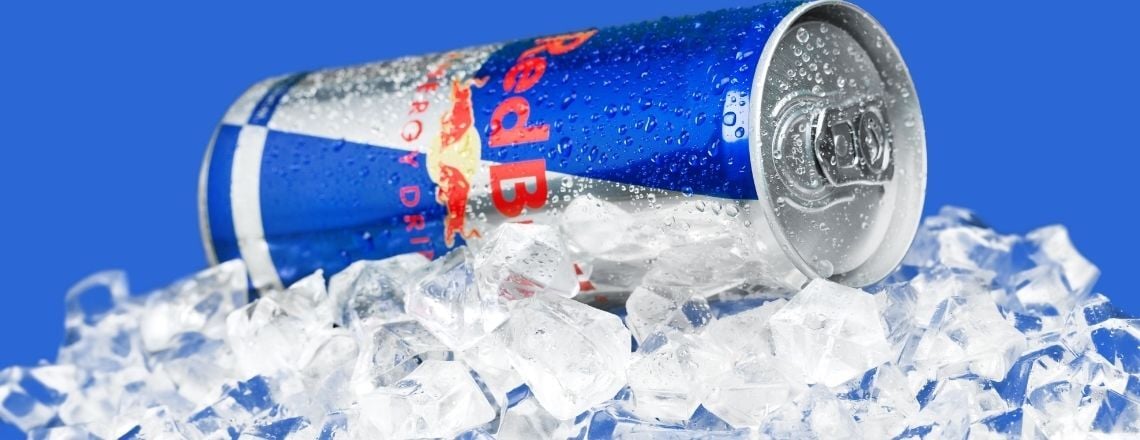 A cold Red Bull energy drink can covered in water droplets resting on a bed of crushed ice against a blue background.