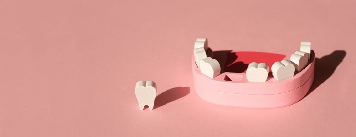 Wooden jaw model representing tooth loss and extraction on a pink background.
