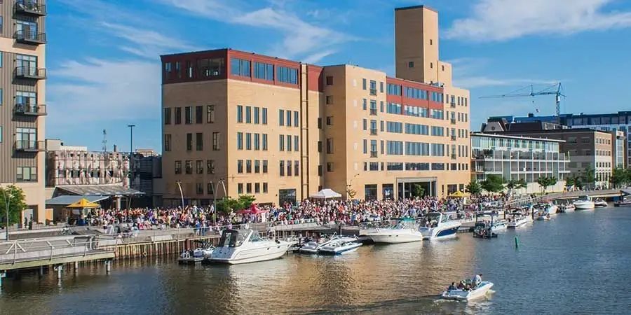 Crowds gather at the CityDeck along the Fox River in Green Bay, Wisconsin, with boats docked on a sunny day.