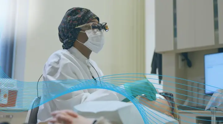A dental hygienist wearing loupes and a face mask examines a patient's teeth.