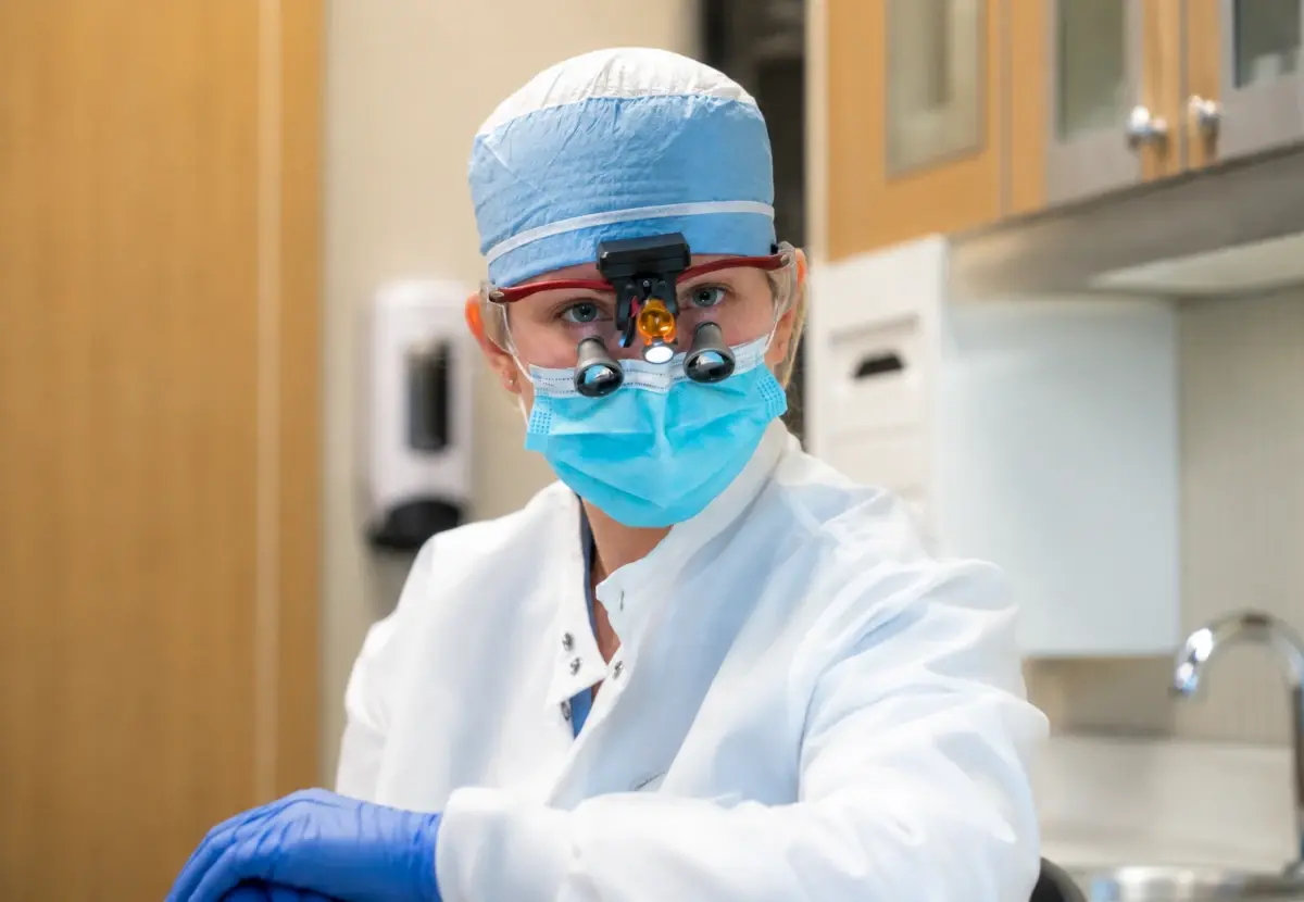 A female dentist wearing magnification loupes, a surgical mask, and gloves in a dental clinic.